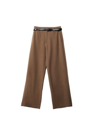 Personne portant une veste taupe zippée avec les manches retroussées et un pantalon marron assorti avec une ceinture noire, une main dans la poche.