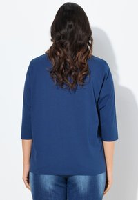 Granatowa, koszula z rękawem o długości trzech czwartych, wykonana z miękkiego materiału, charakteryzująca się luźnym krojem i zaokrąglonym dołem w talii.