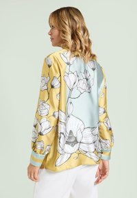 Silk blouse featuring a yellow base with large floral prints in black and white, a blue back panel, and striped cuffs in light blue.