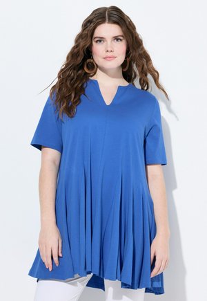 Blaue Tunika mit V-Ausschnitt und kurzen Ärmeln; verfügt über ein gefältetes Design und einen glatten, leichten Stoff; kombiniert mit weißen Hosen.