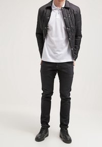 En man klädd i en vit polo, svart vadderad jacka, svarta smala jeans och svarta sneakers står mot en enfärgad ljus bakgrund.