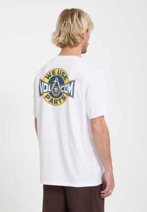Blonde man met de rug naar de camera, draagt een wit T-shirt met een grote gele en blauwe "We Use Volcom Parts '91" afbeelding op de achterkant.