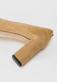 Hnědé semišové lodičky na vysokém podpatku s blokovým podpatkem. Zaoblená špička a černý akcent na podrážce. Hladká textura s bezproblémovým designem.
