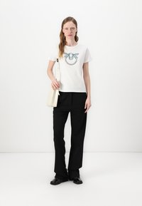 Maglietta bianca a maniche corte con un design grafico blu e turchese; abbinata a pantaloni neri larghi e una borsa a tracolla color crema.