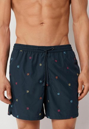 Mann trägt marineblaue Badeshorts mit bunten, bestickten Ankern und einem Tunnelzug in der Taille, steht vor einem neutralen Hintergrund.