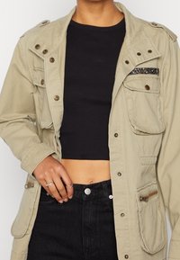 Khaki, lehká bunda s knoflíkovým uzávěrem, velkými kapsami a strukturovanou černou kapsou na hrudi. Černý crop top pod tím.