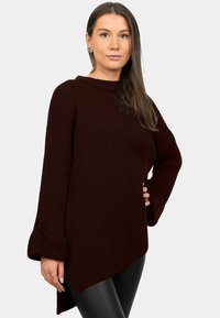 Maglione color borgogna scuro lavorato a maglia con trama a costine, maniche larghe e orlo asimmetrico, abbinato a pantaloni neri aderenti.