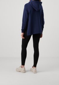 Kvinna som står i svarta leggings, marinblå hoodie och beige sneakers, visad från baksidan mot en enkel ljus bakgrund.