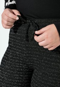 Černé texturované joggery vyrobené z pletené tkaniny s kovovými nitěmi, s pasem na šňůrku držené v rukou.