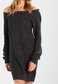 Robe tricotée gris foncé à col bardot avec texture côtelée, manches longues et coupe décontractée. Détail accentué aux poignets.