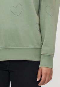 Zeleno svetrovko z majhnimi kovinskimi srčnimi okraski na vsaki strani, z rebrastimi manšetami in gladko teksturo.