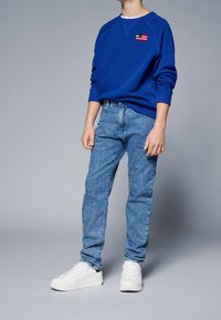 Niebieska bluza z małą czerwoną, białą i niebieską łatą, zestawiona z jasnoniebieskimi dżinsami i białymi sneakersami. Prosty krój i design.