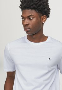 White short-sleeve t-shirt made of soft fabric, featuring a round neckline and a small black logo accent on the chest. Smooth texture.
