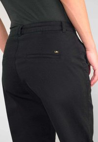 Zwarte broek met een aansluitende pasvorm, met een achterzak en een klein gouden logo-accent. De stof heeft een gladde textuur.