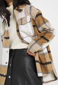 Kvinna i beige polotröja, brun och vit rutig knappjacka, och svart läder kjol med knappar fram, med handen på höften.