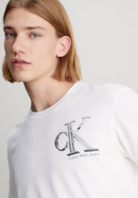 Balta medvilnės marškinėlių su grafiniu logotipu, kuriame yra 'cK' ir 'Calvin Klein Jeans' pilkos spalvos. Dizainas yra paprastas, su apykakle.