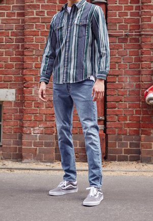 Personne portant une chemise rayée verticalement verte et bleu marine, un jean bleu et des baskets grises, debout sur l'asphalte devant un mur en briques rouges.