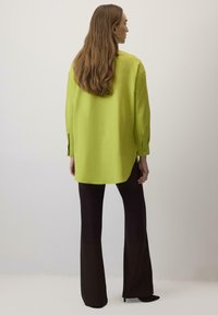 Camicia oversize verde lime con maniche lunghe e polsini con bottoni, abbinata a pantaloni neri a campana. Texture liscia, con design a orlo irregolare.