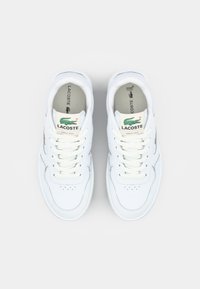 Witte leren sneakers met vetersluiting, geperforeerd teenontwerp en groen Lacoste-logo op de tong en zijkant. Zichtbare gevoerde binnenzool.