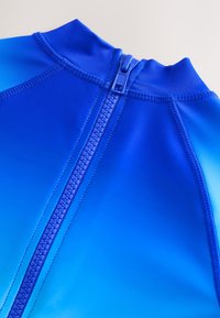 Muta da sub azzurra con cerniera frontale, texture liscia e sfumatura dal blu scuro al chiaro. Dettagli delle cuciture visibili sulle spalle.