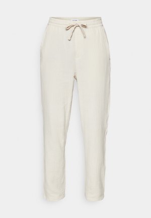 Calças de algodão texturizadas, em bege claro, com cintura elástica e cordão. Apresentam bolsos laterais e pernas ajustadas.