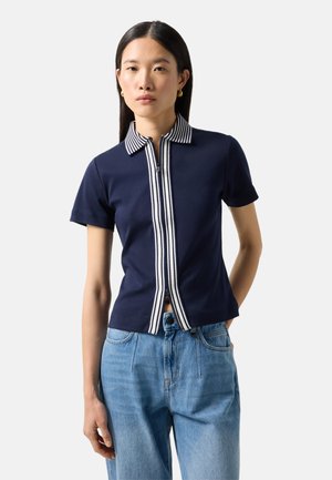 Junge Frau mit glatten schwarzen Haaren, die ein dunkelblaues kurzärmeliges Poloshirt mit weiß gestreiftem Kragen und Reißverschluss sowie hellblaue Jeans trägt.