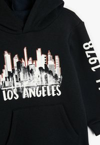 La sudadera negra presenta un gráfico de un horizonte urbano con "LOS ÁNGELES" impreso en blanco. Incluye un bolsillo frontal y una capucha.