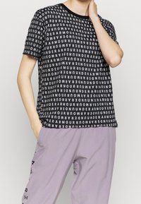 Camiseta negra de manga corta con un patrón repetido del logo blanco de "Under Armour". Cuello redondo, combinada con pantalones de color púrpura claro.