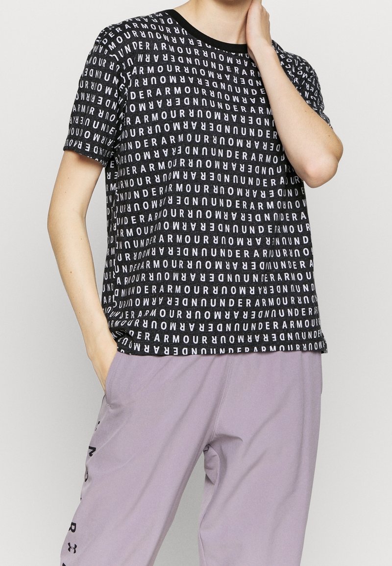 Camiseta negra de manga corta con un patrón repetido del logo blanco de "Under Armour". Cuello redondo, combinada con pantalones de color púrpura claro.