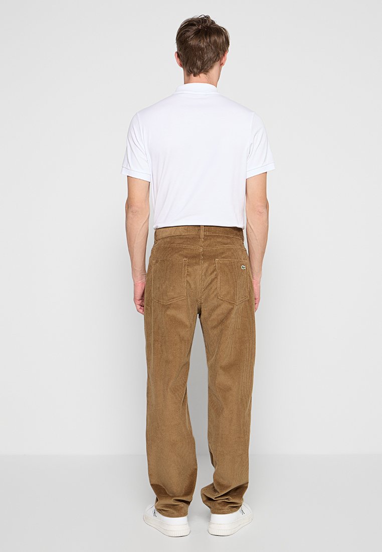 Pantalon en velours côtelé marron coupe droite, avec poches arrière et un détail de logo. Associé à un polo blanc uni et des chaussures blanches.