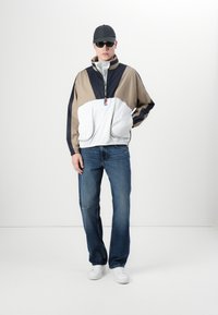 Fargeblokket vindjakke i beige, marineblå og hvit, med høy krage og glidelås; kombinert med blå jeans og hvite sneakers.