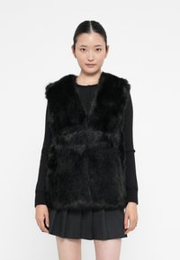 Junge Frau, die eine schwarze Fleece-Weste über einem langärmligen schwarzen Oberteil und einem gefalteten schwarzen Rock trägt, steht vor einem weißen Hintergrund.