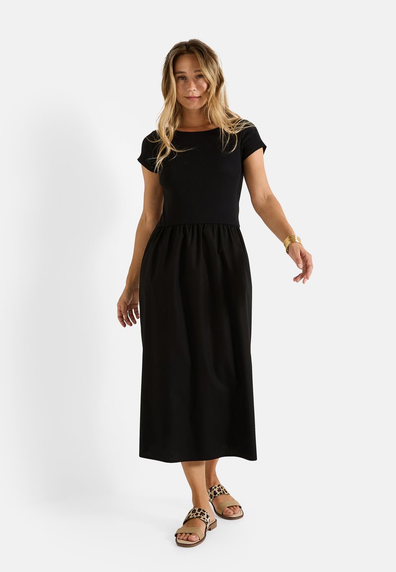 Robe noire avec un haut ajusté et une jupe froncée ; associée à des sandales à imprimé léopard. Texture lisse, manches courtes et design longueur cheville.