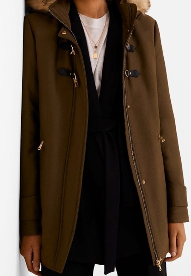 Femme portant un manteau marron à capuche avec bordure en fourrure, couche intérieure noire, chemise blanche et deux colliers en or, debout devant un fond blanc.