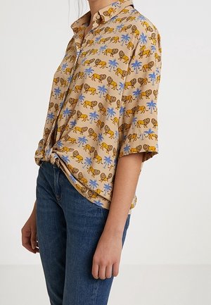 Kurzärmliges Hemd in Beige mit Löwen- und Palmenmuster, getragen mit blauen Jeans an einer stehenden Person.