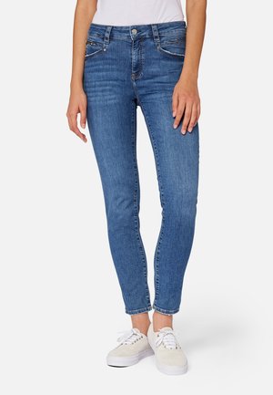 Femme portant un jean bleu slim taille mi-haute et des baskets blanches, debout devant un fond uni.