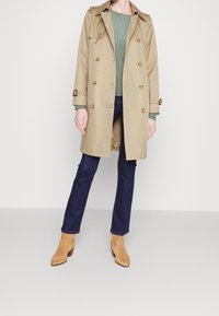 Beige trenchcoat med guldknappar, bälte i midjan och axelklaffar. Bärs över en grön topp, kombinerad med mörkblå jeans och bruna ankelstövlar.