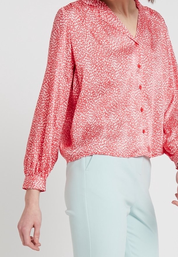 Blusa rossa a motivi con maniche lunghe, colletto e chiusura a bottoni. Il tessuto appare morbido e leggero, abbinato a pantaloni azzurri chiari.
