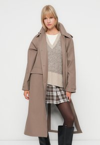 Long beige trench coat over a grey sweater and plaid skirt. Black knee-high boots complete the outfit, with a smooth fabric texture.
