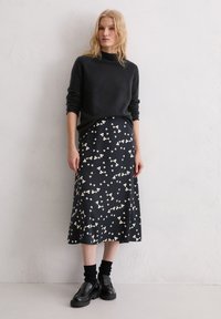 Schwarzer Pullover mit hohem Kragen, kombiniert mit einem marineblauen Rock mit weißen Blumenmustern. Das Modell trägt schwarze Schuhe und knielange Socken.