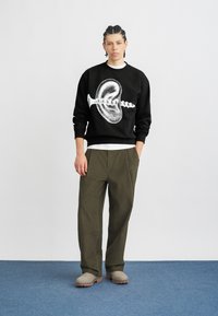 Μαύρο φούτερ με λευκό γραφικό σχέδιο, συνδυασμένο με χαλαρές ελιάς πράσινες τσόχες και γκρι παντόφλες. Καθαρή, μινιμαλιστική αισθητική.