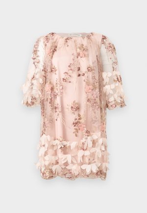 Femme portant une robe rose courte et transparente avec des applications florales 3D et de la broderie, associée à des talons ouverts noirs à bride à la cheville.