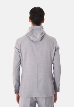 Uomo con giacca da abito grigio chiaro con cappuccio attaccato, visto da dietro su sfondo bianco.