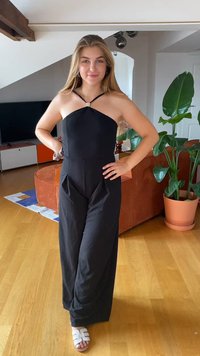 Mono negro con diseño de cuello halter, acentuado por un detalle de strass. Presenta piernas anchas y tejido texturizado. Persona vestida con sandalias blancas.