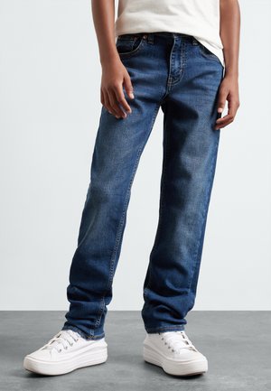 Jean en denim bleu foncé avec une coupe décontractée, présentant des accents de couture, cinq poches et une texture usée, associés à des baskets blanches.