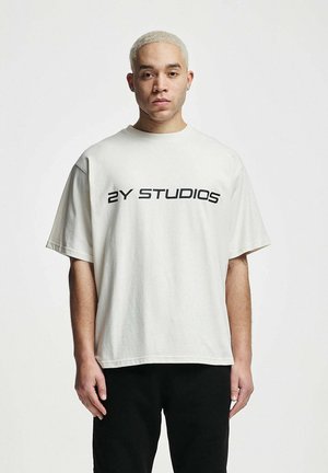 Kurzes, beiges, übergroßes T-Shirt aus Baumwolle, mit einem schwarzen Logo "2Y STUDIOS" quer über die Brust in fetter Schrift gedruckt.