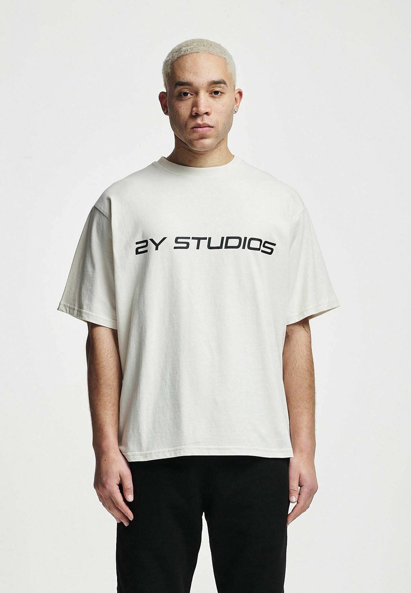 Lyhyt vaalean beige ylisuuri t-paita, joka on valmistettu puuvillasta. Paidassa on musta logo "2Y STUDIOS" tulostettuna rinnan yli rohkealla fontilla.