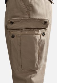 Beżowe spodnie cargo charakteryzują się dużą, otwartą kieszenią z zapięciem na guziki oraz teksturowaną tkaniną, zaprojektowaną z myślą o funkcjonalności i wytrzymałości.