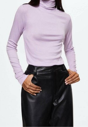 Persona que lleva un top de manga larga con cuello alto de color púrpura claro, metido dentro de unos pantalones de cuero negros de cintura alta, con las manos en los bolsillos.