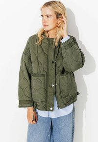 Veste matelassée vert olive avec un col rond, fermeture à pression et deux poches latérales, portée par-dessus une chemise rayée bleu et blanc.